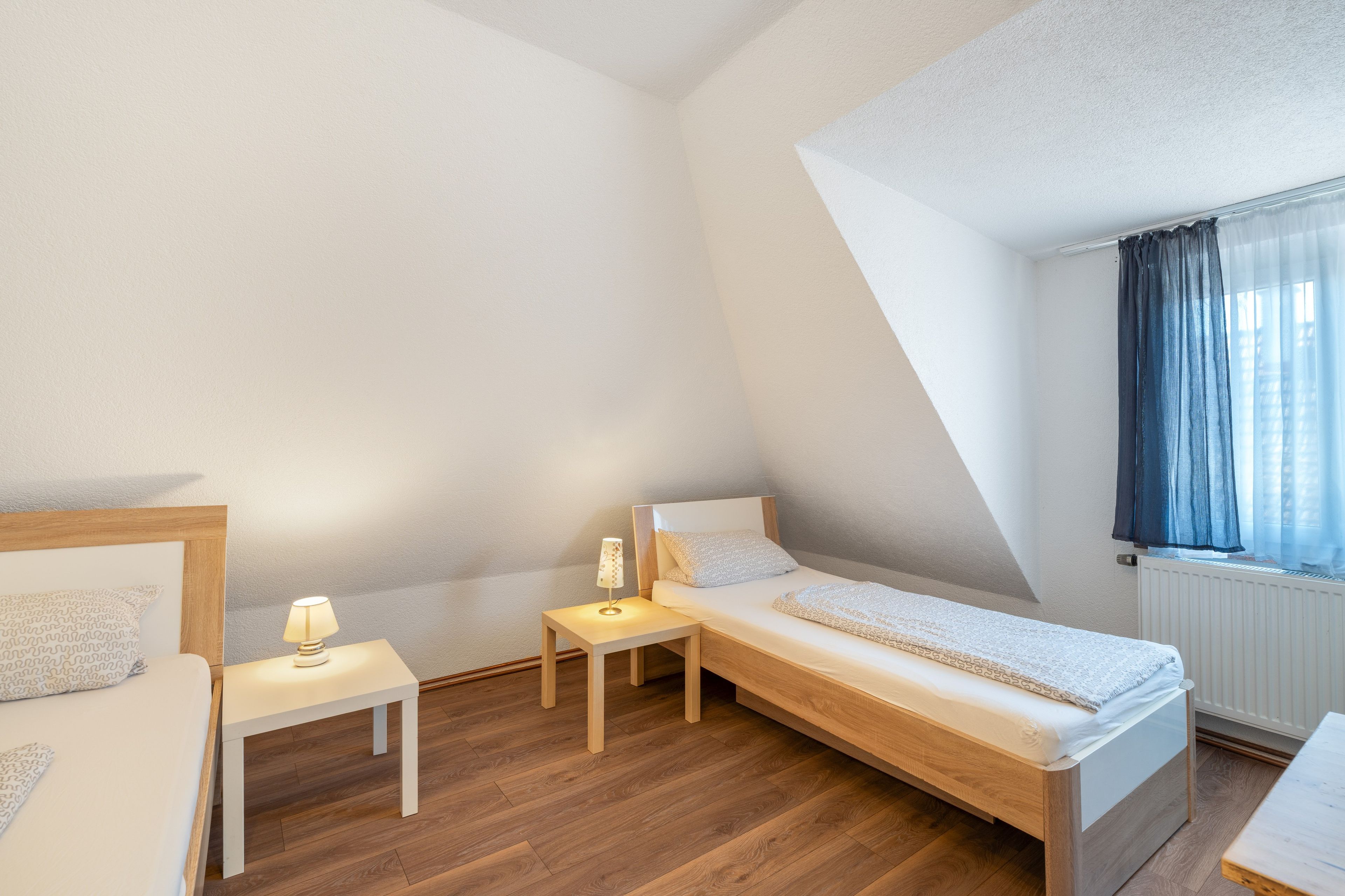 A small, well-lit bedroom with a single bed, a bedside table with a lamp, and a window with curtains.