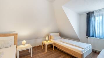A small, well-lit bedroom with a single bed, a bedside table with a lamp, and a window with curtains.