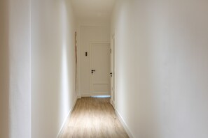 Un couloir étroit, doté d’un plancher en bois et de murs blancs, qui mène à une porte fermée sur laquelle est apposée une plaque avec un numéro.