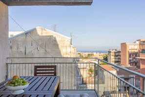 Ein Balkon mit einem Tisch, auf dem sich eine Obstschale befindet, mit Blick auf eine Stadtlandschaft mit Gebäuden und blauem Himmel.