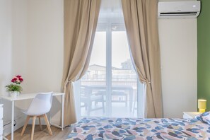 A hotel room with a bed, a desk, a chair, a window with curtains, and an air conditioning unit. - Apartment 'Room 002' with Sea View, Wi-Fi, and Air Conditioning (Catania)