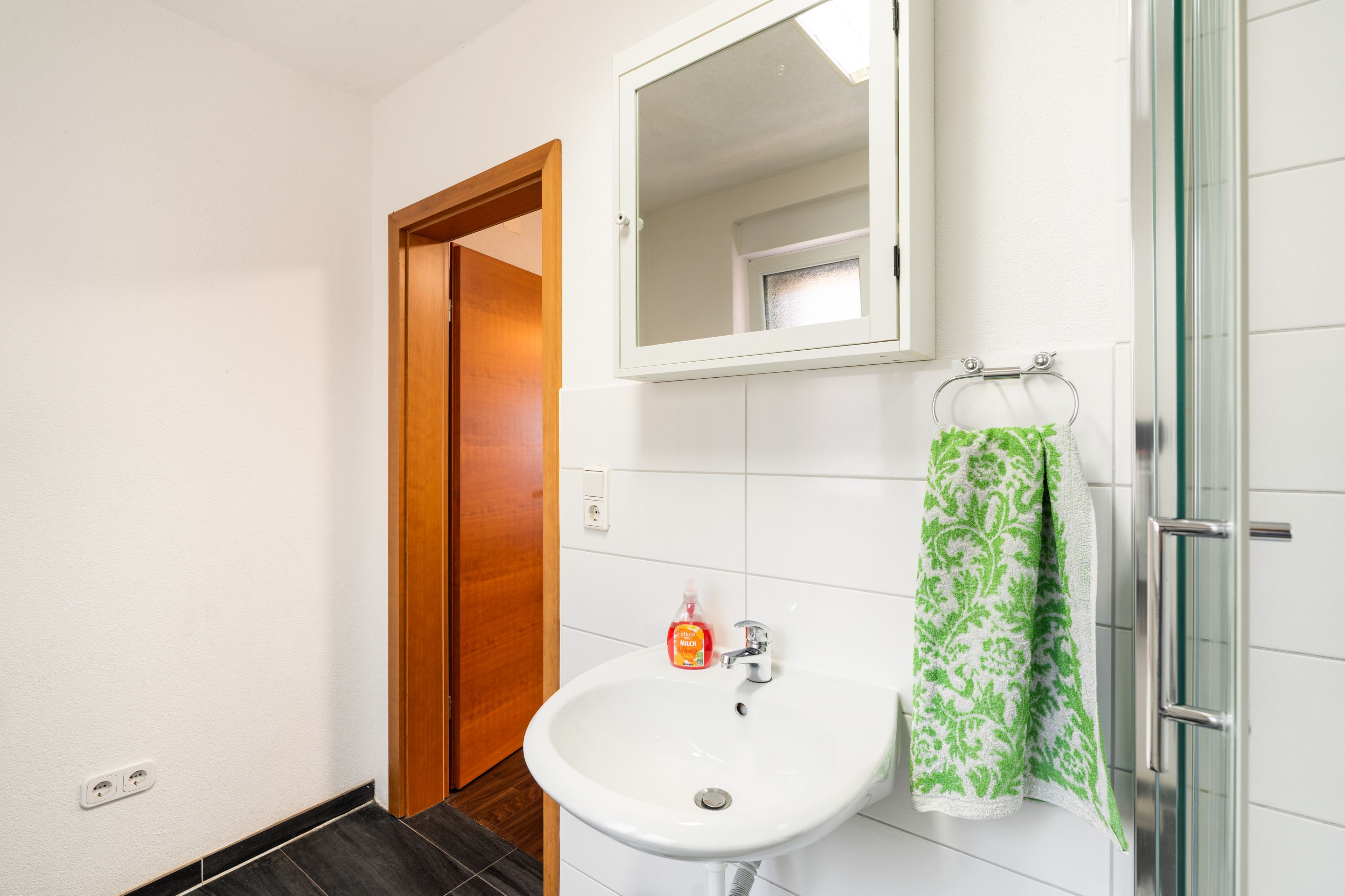 A bathroom with a white sink, a mirror, a glass shower enclosure, and a green patterned towel.