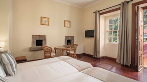 Ein Hotelzimmer mit einem großen Bett, zwei Korbstühlen, einem kleinen Tisch, einem Kamin, einem Fernseher und einem Fenster mit Vorhängen.