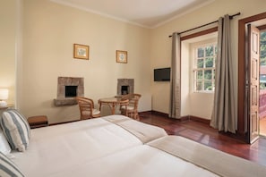 A hotel room with a large bed, two wicker chairs, a small table, a fireplace, a TV, and a window with curtains. - Vrbo Property (Santana)