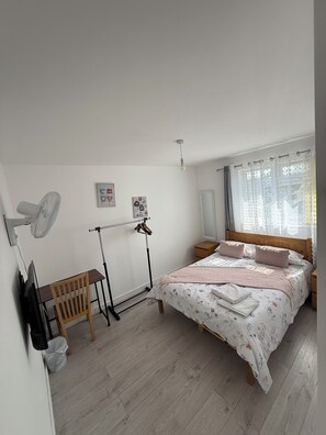Quarto com cama, escrivaninha, cadeira, ventoinha e janela com cortinas.