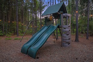 A playground with a slide and climbing structures in a wooded area. - 4 King Suites | Hot Tub & Game Room (Broken Bow)