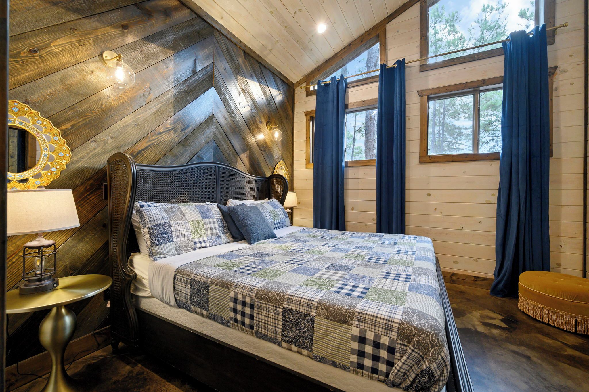 A bedroom with a wooden sloped ceiling, a bed with a quilt, a nightstand, a lamp, and a window with curtains.