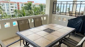 A balcony with a table and chairs, overlooking a residential area with palm trees and buildings. - Cozy 2BD ocean-front condo with tennis courts (Las Jarretaderas)