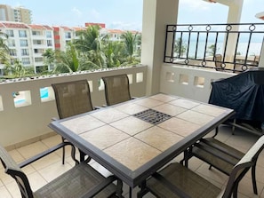 A balcony with a table and chairs, overlooking a residential area with palm trees and buildings. - Cozy 2BD ocean-front condo with tennis courts (Las Jarretaderas)
