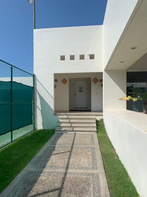 A modern building entrance with a concrete pathway leading to a door, a green fence on one side, and a clear sky. - Cozy 2BD ocean-front condo with tennis courts (Las Jarretaderas)