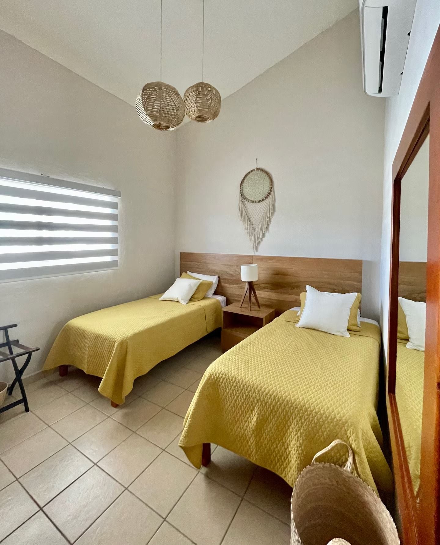 Une chambre d’hôtel avec deux lits, une table de chevet, une lampe, un attrape-rêves et une fenêtre avec des stores.