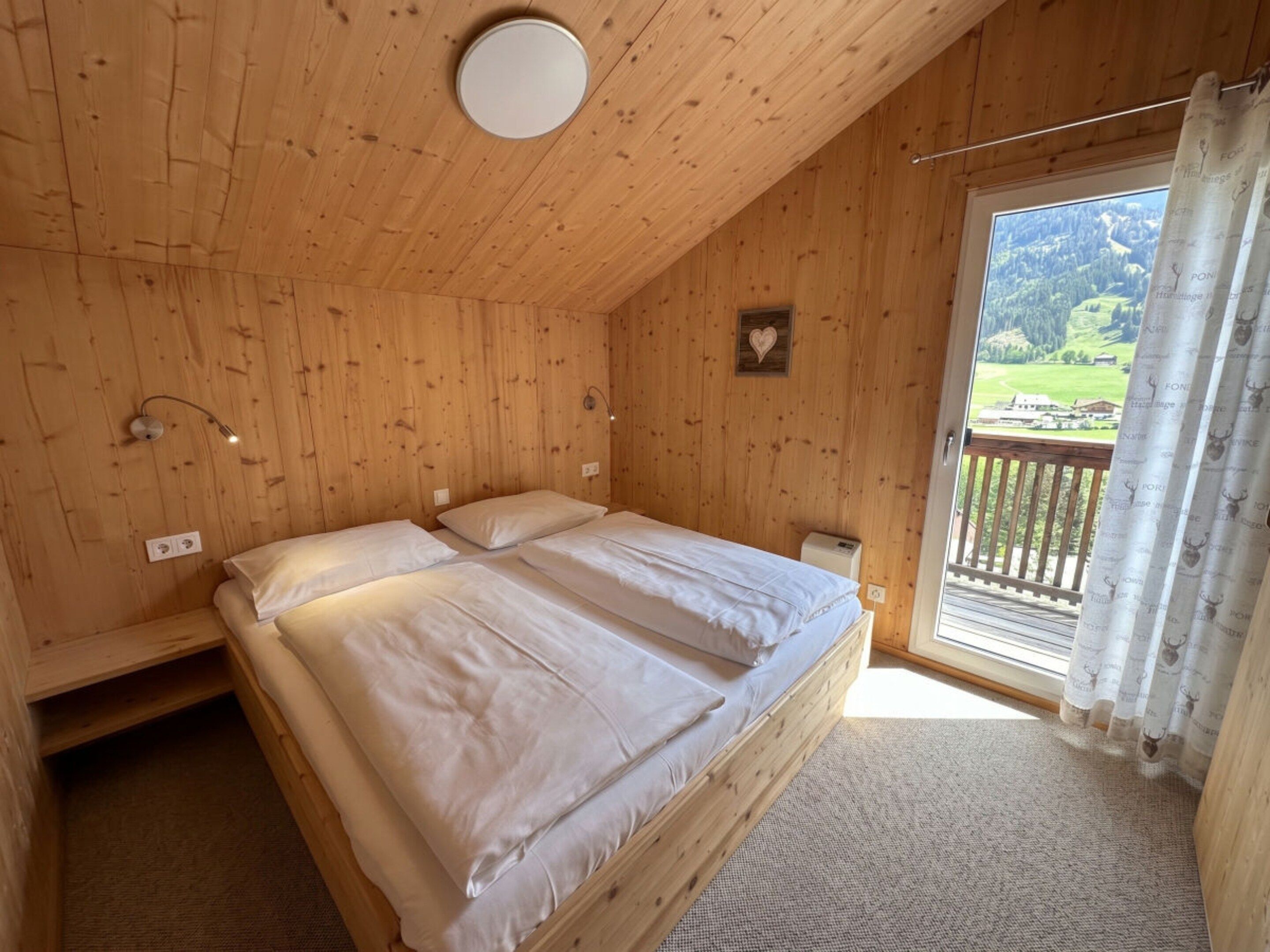 Quarto de cabana de madeira com duas camas, uma luminária no teto, janela com cortinas e vista para uma paisagem verde.