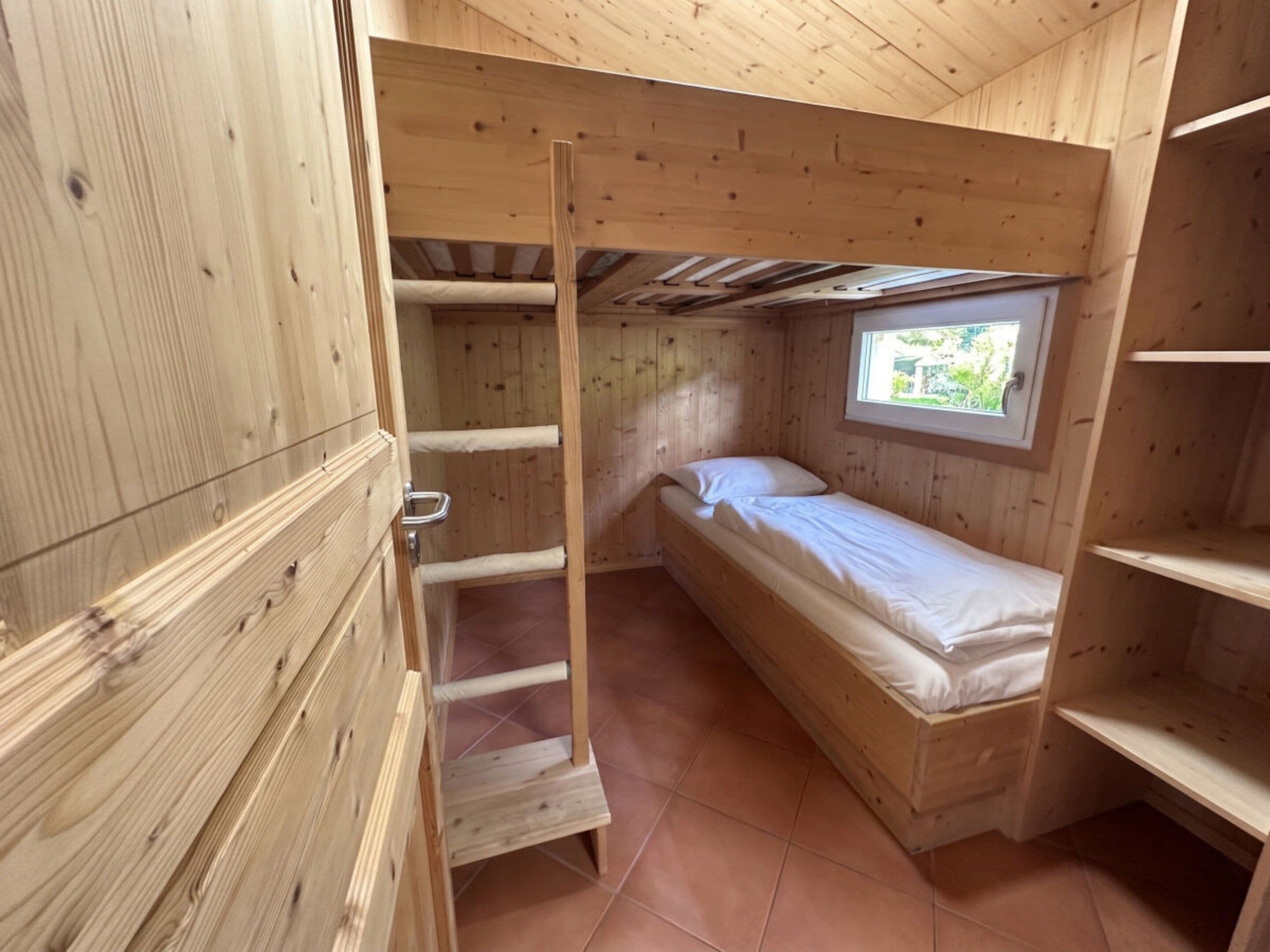 A small wooden room with a single bed, a window, and a wooden ladder.