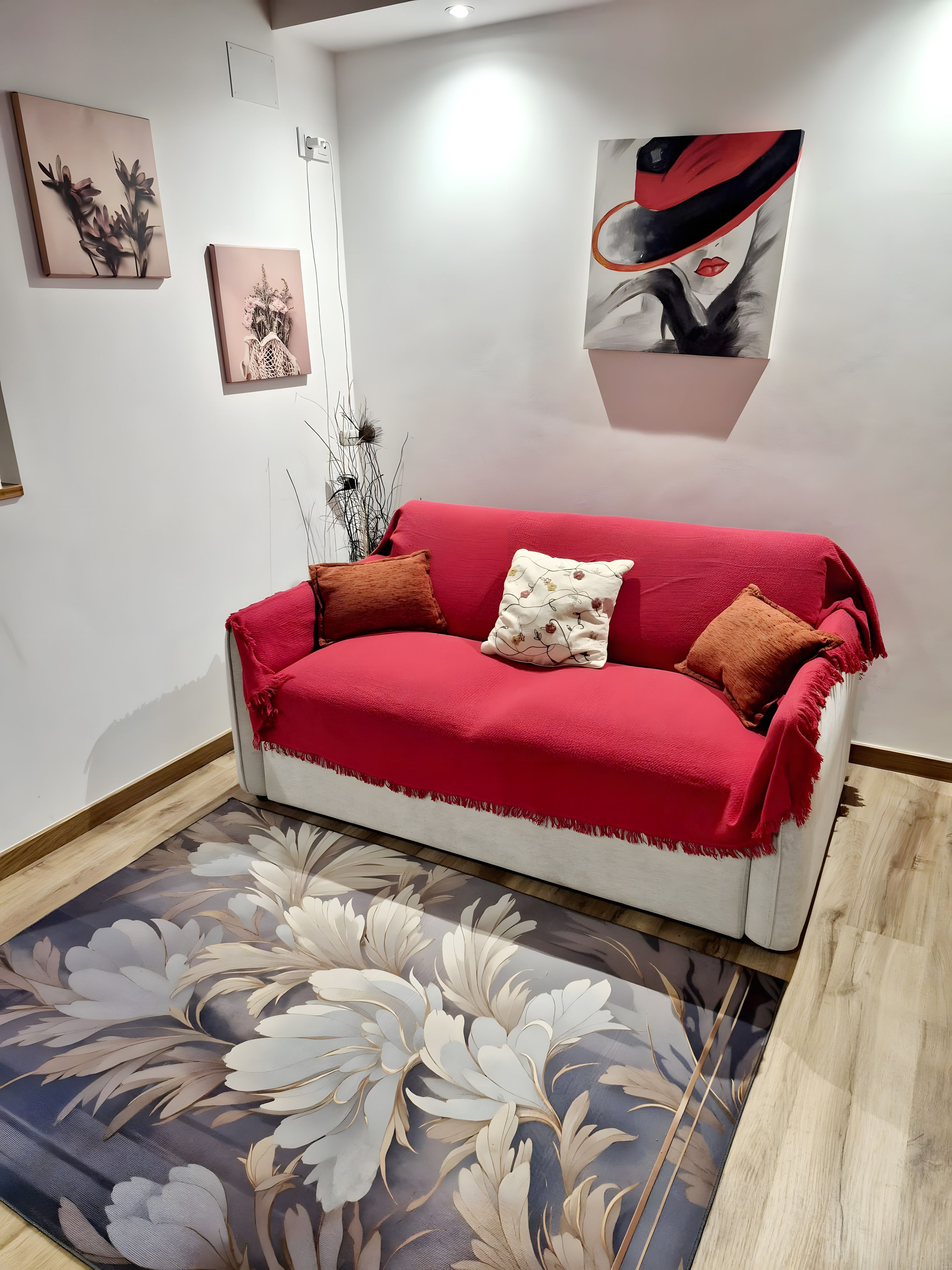 Un sofá rojo con dos almohadas, una alfombra floral y una obra de arte en la pared.