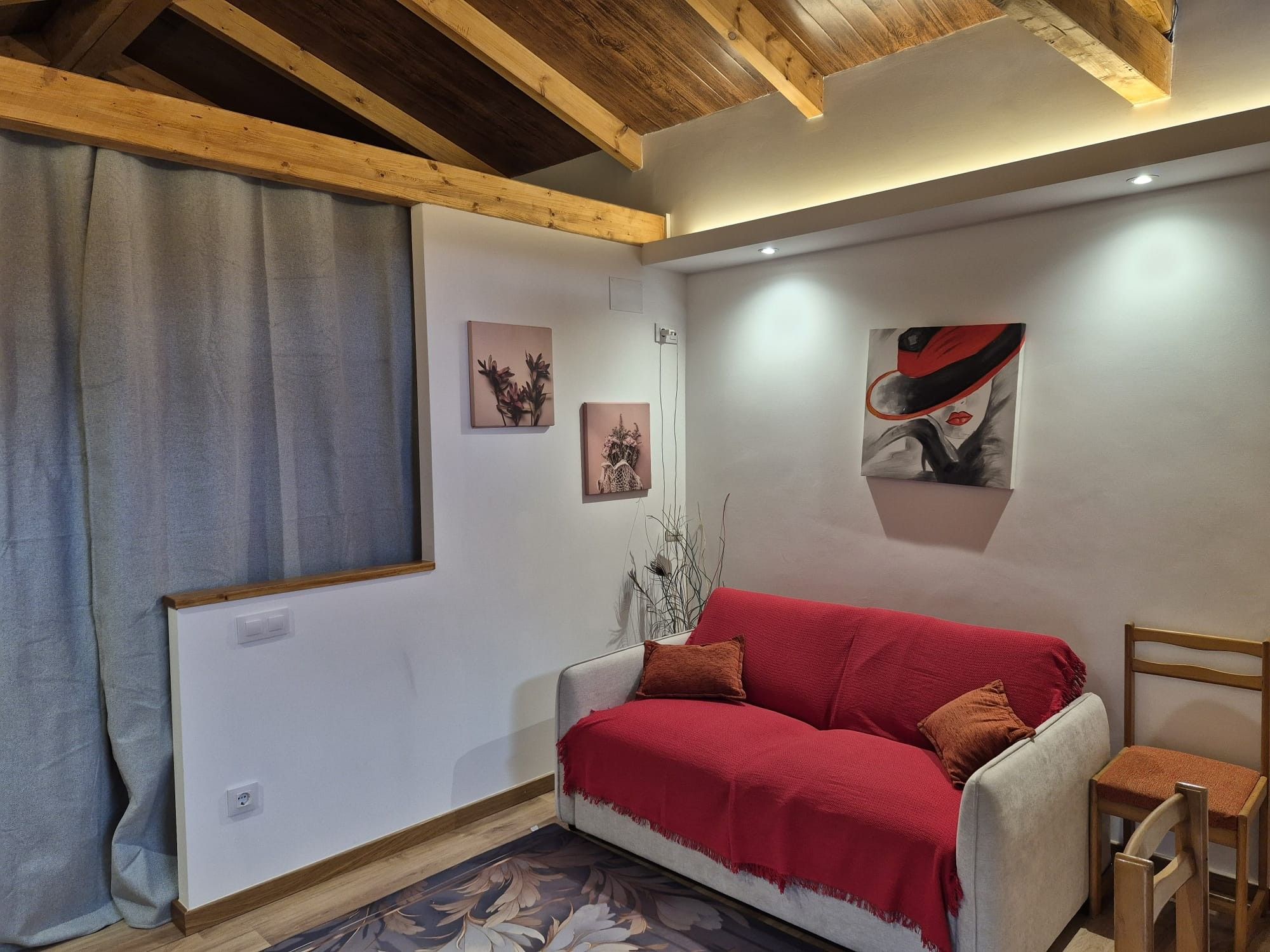 Een knus vertrek met een houten plafond, een rode bank, een klein houten stoeltje en twee schilderijen aan de muur.