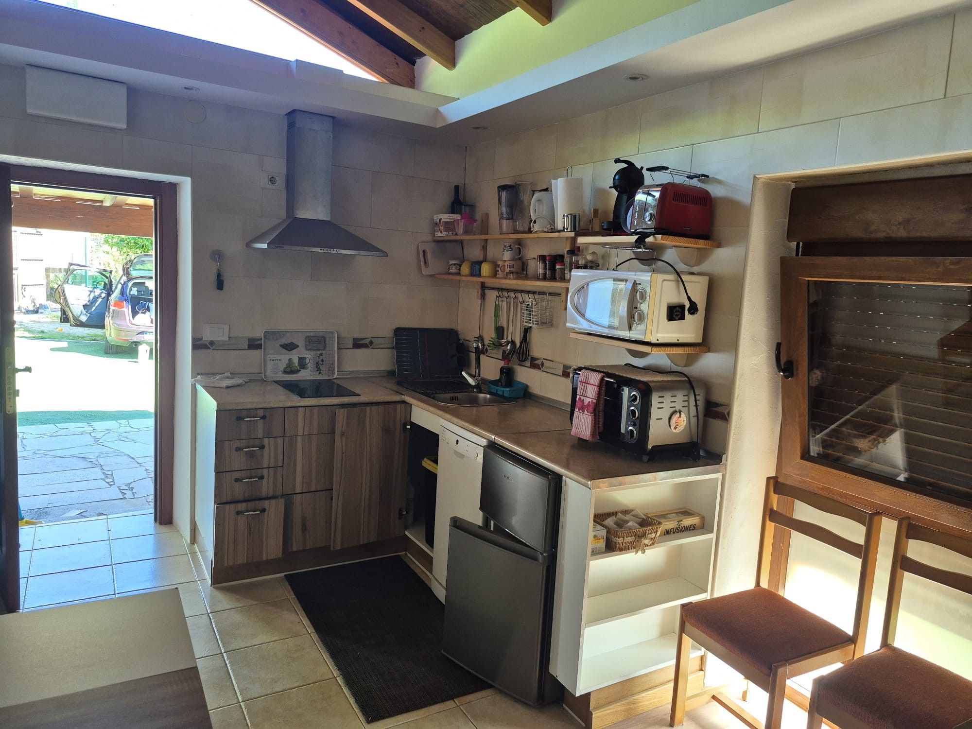 Cocina compacta equipada con lavavajillas, microondas y tostadora. Se observa un armario de madera con cajones y una pequeña zona de estar con una silla.