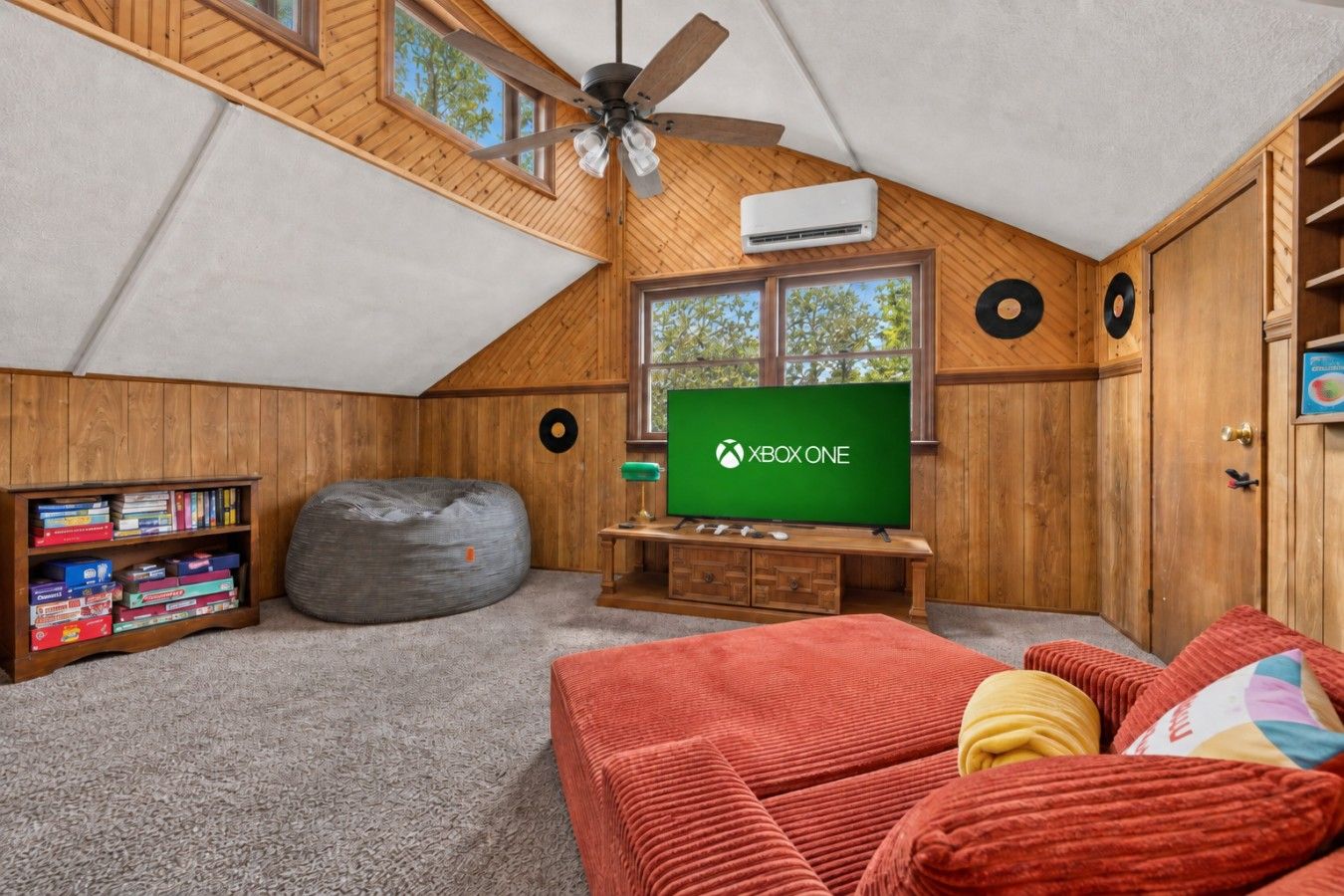 Ein gemütliches Zimmer mit schrägen Wänden, Holzwänden, einem Deckenventilator, einer Klimaanlage, einem großen Fenster, einem Sitzsack, einem Bücherregal, einem Fernseher und einem roten Sofa.