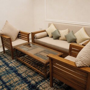 A living room with a sofa, two wooden chairs, a glass coffee table, and a patterned rug. - Kamdhenu Lok (Haridwar)