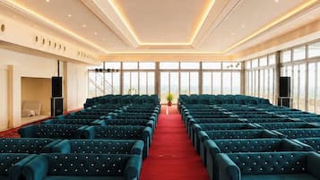 Een congreszaal met rijen zitplaatsen, een rood tapijt en grote ramen.