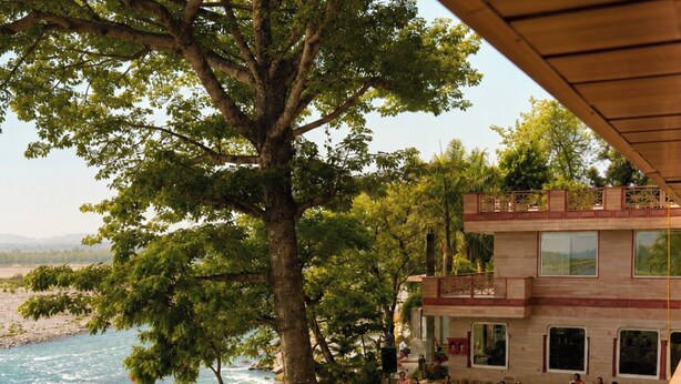 An outdoor event with a river view, a large tree, and a building with multiple windows. - Kamdhenu Lok (Haridwar)