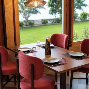 A dining table set with plates, glasses, and a bottle, surrounded by chairs, with a view of a garden outside.