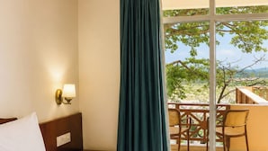 A hotel room with a bed, a nightstand with a lamp, a window with a view of the outdoors, and a balcony with chairs. - Kamdhenu Lok (Haridwar)