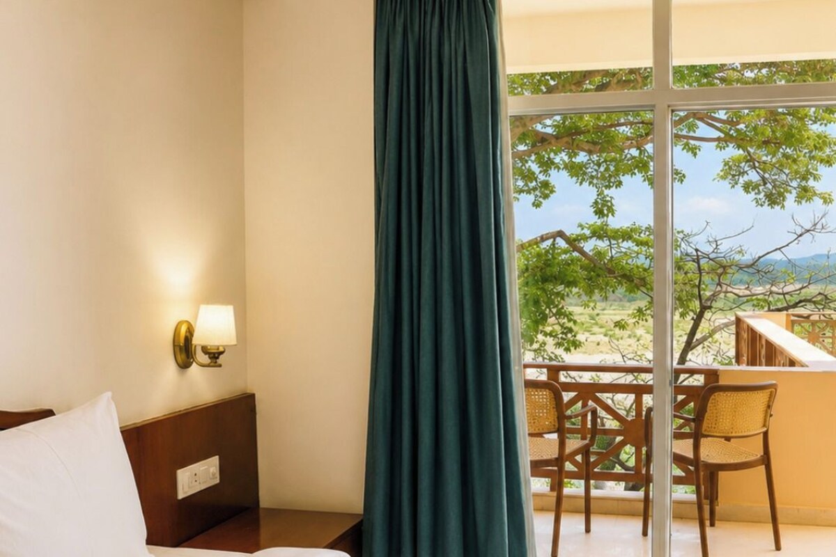 A hotel room with a bed, a nightstand with a lamp, a window with a view of the outdoors, and a balcony with chairs.