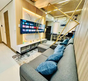 A modern living room with a large flat-screen TV mounted on a wall, a grey sofa with blue cushions, and a wooden slatted ceiling design. - Royal Fame Residence (Mandaluyong)