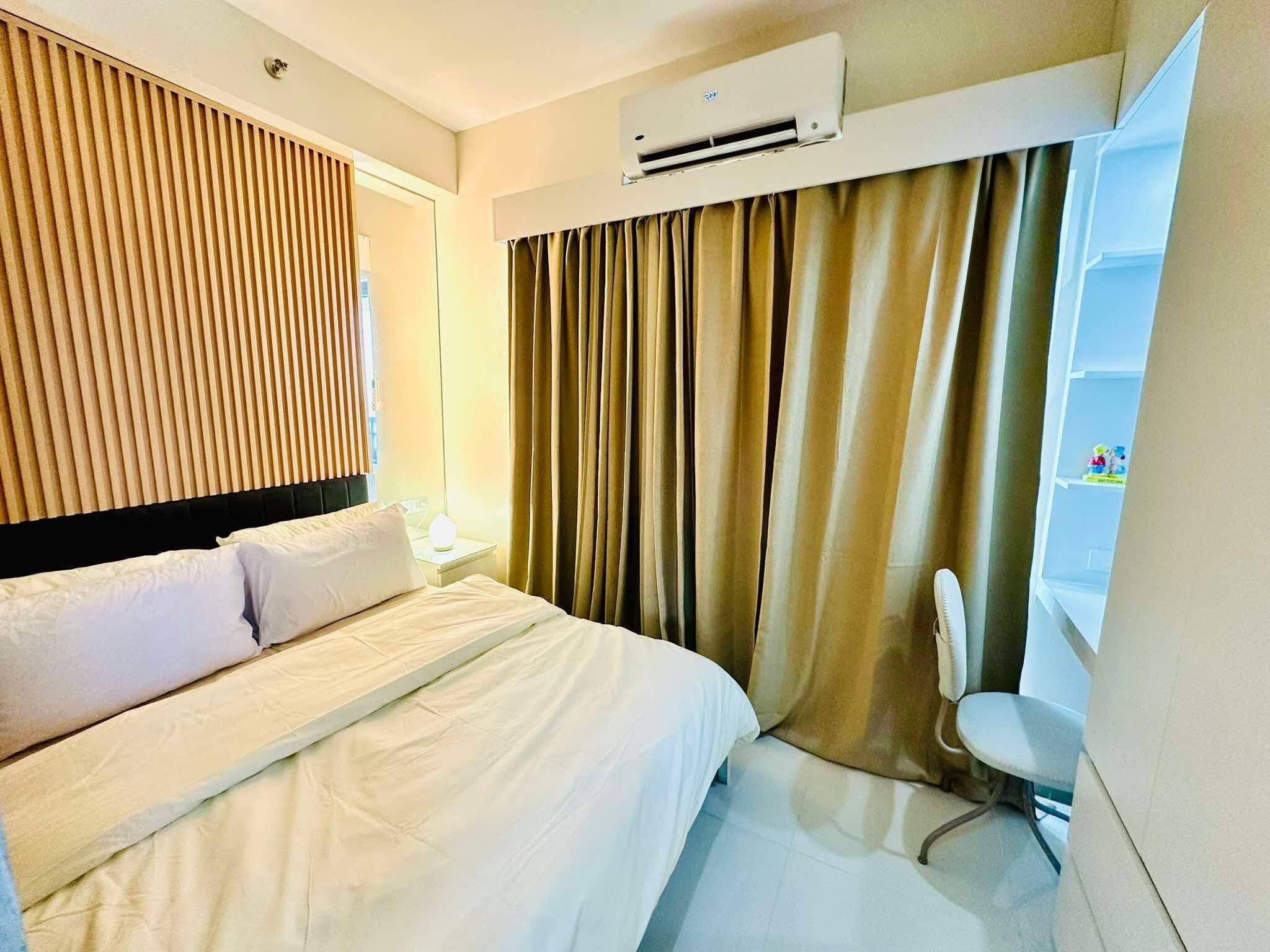A hotel room with a bed, a chair, a desk, a shelf with items, and an air conditioning unit.