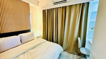 A hotel room with a bed, a chair, a desk, a shelf with items, and an air conditioning unit.