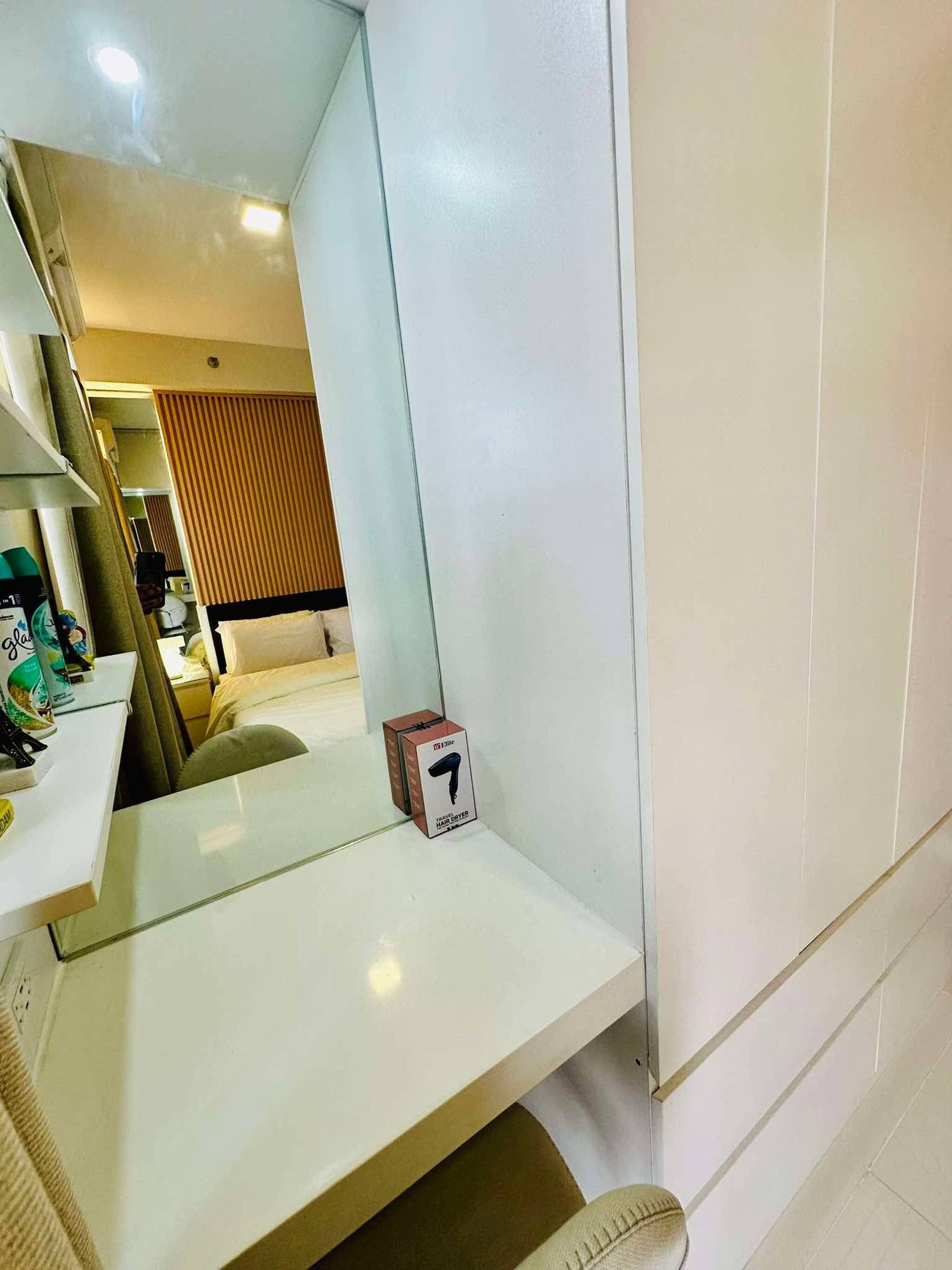 A hotel room with a white bed, a small table with a box, and a mirror reflecting the room.
