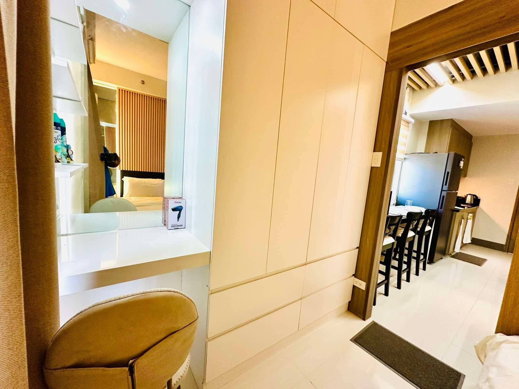 A hotel room with a bed, a chair, a desk, a mirror, and a kitchenette.