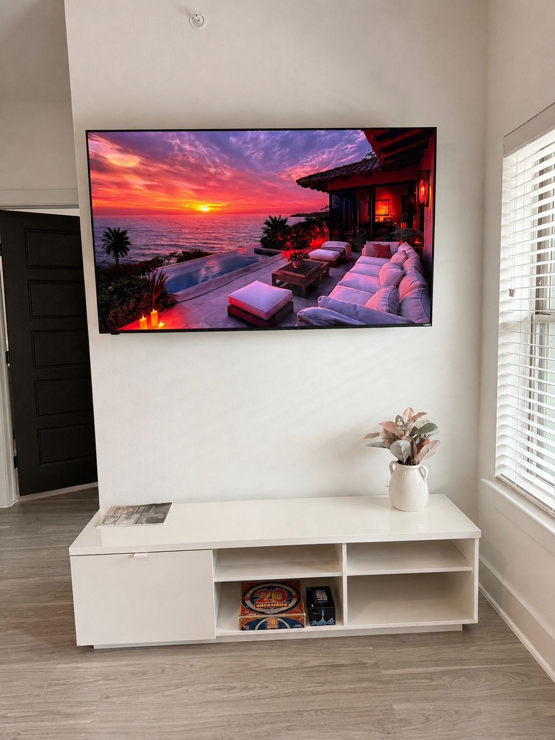 Un téléviseur à écran plat fixé au mur, un meuble de rangement blanc en dessous, et une fenêtre avec des stores sur le côté droit.