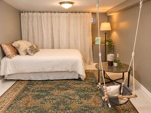 Quarto com cama, cadeira suspensa, luminária e pequena mesa com planta.
