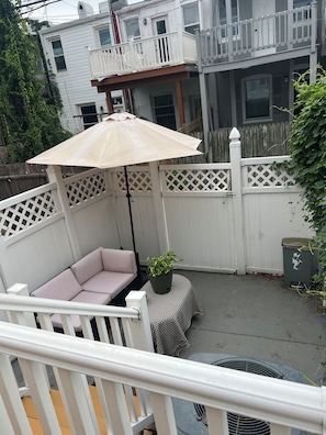 Une terrasse avec un coin salon agrémenté de coussins, un pot de fleurs et une poubelle.