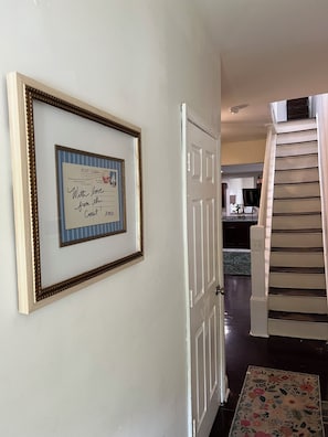 Un couloir avec une carte postale encadrée accrochée au mur, une porte blanche et un escalier recouvert d’un tapis à motifs.