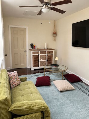 Una sala de estar con un sofá verde, una mesa de centro de vidrio, un ventilador de techo, un televisor montado en la pared y una alfombra estampada.