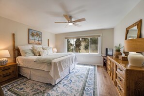 Un dormitorio con cama, mesitas de noche, ventilador de techo, ventana grande y vista a palmeras.