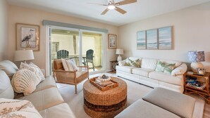 A living room with a ceiling fan, a glass door leading to an outdoor area, a white sofa, a wicker ottoman, and a side table with a lamp and books. - 2-bedroom Condo in St. Augustine- Private Beach Beach Access + Pool! (St. Augustine)