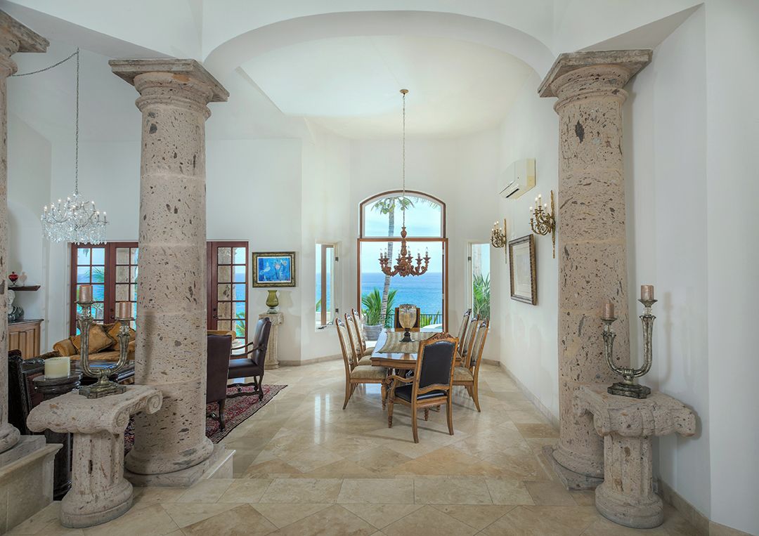 A spacious dining area with a view of the ocean, featuring a large table and chairs, stone columns, and a chandelier.