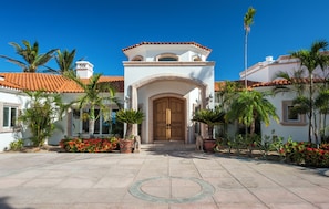 A building with a central entrance, flanked by palm trees and red flowers, under a clear blue sky. - Villa Paraiso Ocean front 8-bedroom Villa in Palmilla Sur Private beach (Palmilla)
