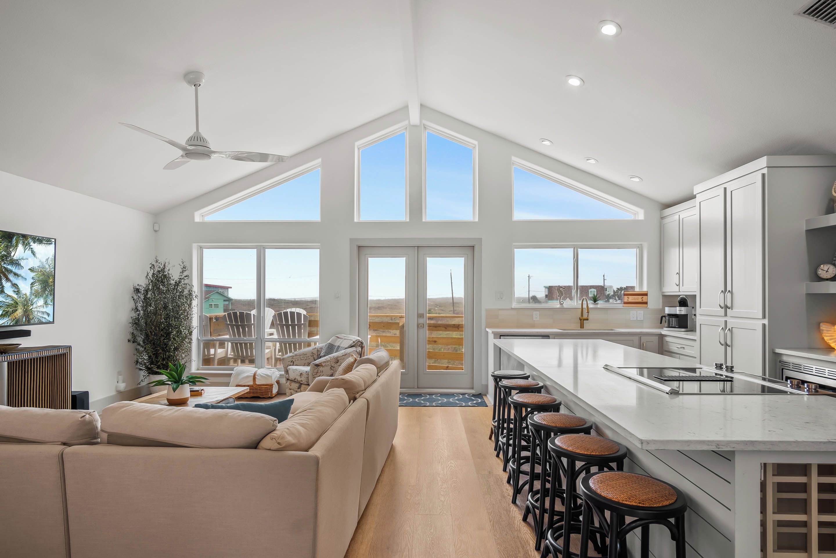 A spacious living area with a high ceiling, large windows, a ceiling fan, and a kitchen with a bar area.