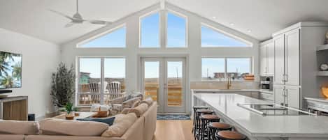 A spacious living area with a high ceiling, large windows, a ceiling fan, and a kitchen with a bar area.