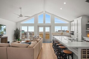 A spacious living area with a high ceiling, large windows, a ceiling fan, and a kitchen with a bar area. - Tan Lines and Fine Wines (Port Aransas)