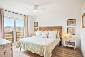 A bedroom with a bed, bedside tables, a ceiling fan, and artwork on the walls. - Tan Lines and Fine Wines (Port Aransas)