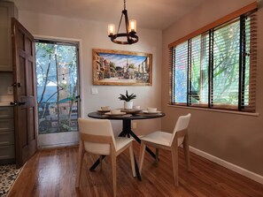 A dining area with a round table, two chairs, a chandelier, and a window with blinds. - Comfy Courtyard 1 Bedroom at Jackson Market (Culver City)