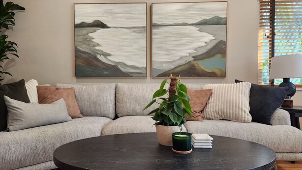 Ein Wohnzimmer mit grauem Sofa, zwei Gemälden, einem runden schwarzen Tisch und einer Topfpflanze.