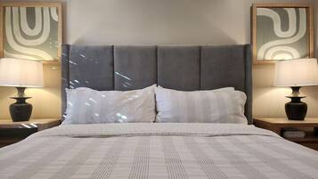 A bed with a grey upholstered headboard, two bedside lamps, and two abstract paintings on the walls.