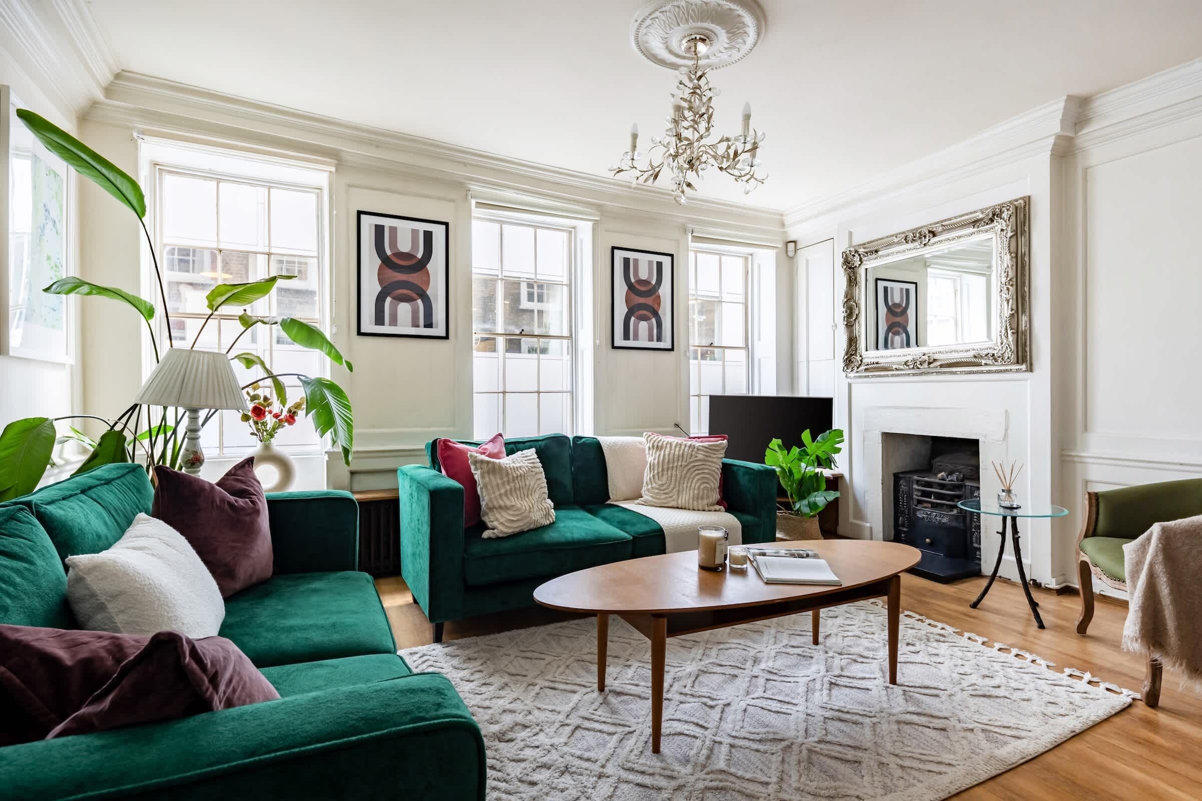 A living room with a green sofa, a coffee table, and a fireplace. There are framed pictures on the wall and a chandelier above the fireplace.