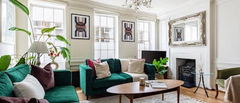 A living room with a green sofa, a coffee table, and a fireplace. There are framed pictures on the wall and a chandelier above the fireplace.